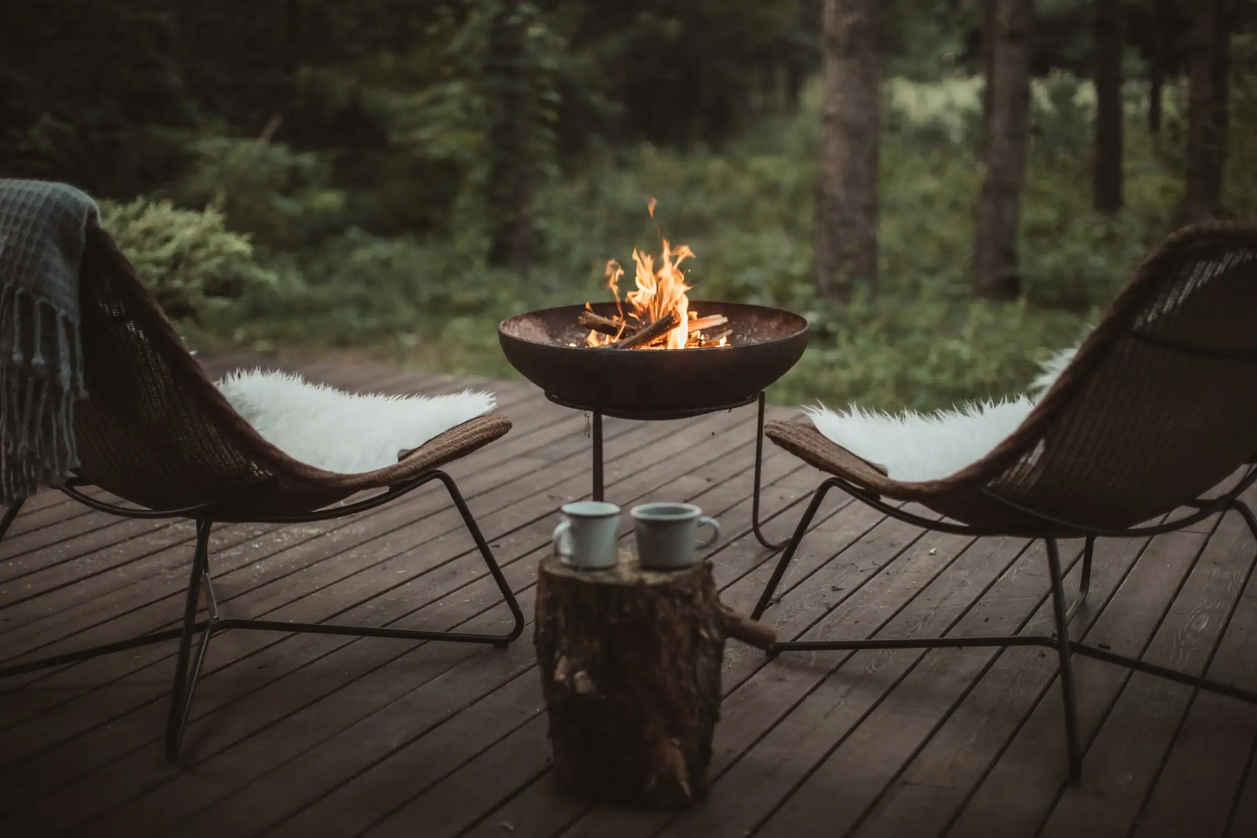 Feu de camp crépitant entre deux chaises sur une terrasse en bois, entourée d'arbres verts.